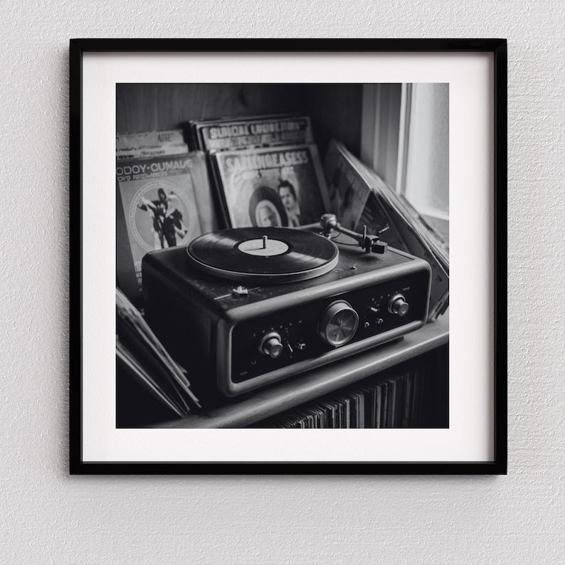 May include: Black and white photograph of a vintage record player with a vinyl record on the turntable. The record player is on a shelf with several vinyl records in the background. The image is framed in a black square frame.