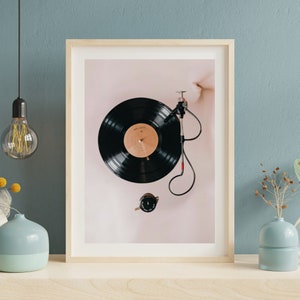 May include: A framed print of a black vinyl record on a turntable with a pink background. The record is playing and the needle is on the record. The turntable is a simple one with a black arm and a silver base.