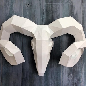 May include: White papercraft ram skull with large, curved horns. The horns are made of folded paper and have a geometric design. The skull is mounted on a dark blue wooden background.