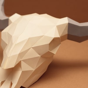 May include: A papercraft bull skull with gray horns. The skull is made of white paper and has a geometric design.