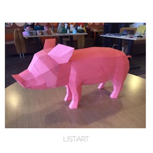 May include: A pink, geometric paper pig sculpture. The pig is constructed from multiple flat, angular planes, giving it a polygonal appearance. The sculpture is displayed on a wooden table. The word "LISTART" is printed at the bottom.