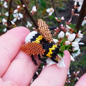 Peut inclure: Un bourdon en perles avec des perles noires, jaunes et brunes. Le bourdon est assis sur la main d'une personne devant une fleur blanche.