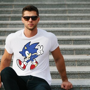 May include: A white t-shirt with a blue and red cartoon character wearing red shoes and a blue headband. The character is holding up two fingers in a peace sign. The character is Sonic the Hedgehog.