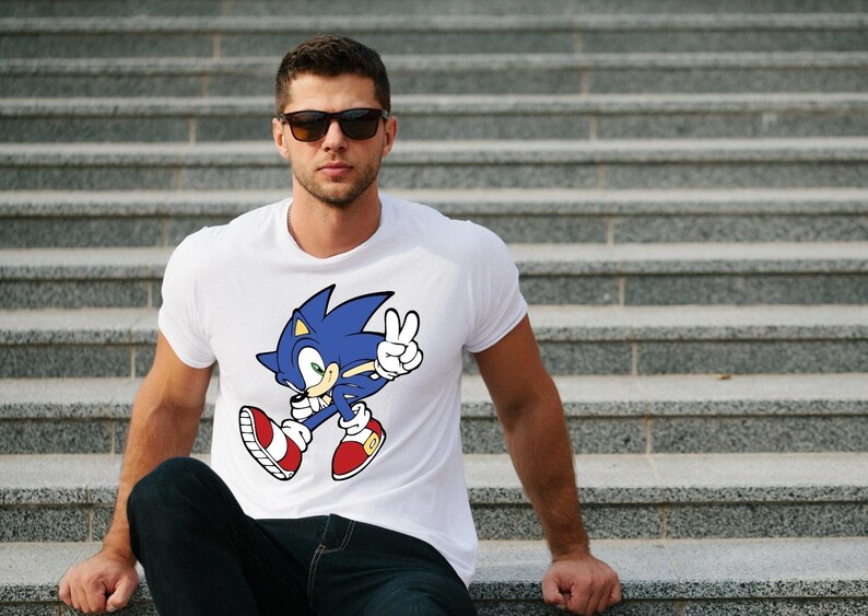 May include: A white t-shirt with a blue and red cartoon character wearing red shoes and a blue headband. The character is holding up two fingers in a peace sign. The character is Sonic the Hedgehog.