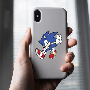 May include: A white phone case with a blue and red Sonic the Hedgehog sticker. Sonic is running and giving a peace sign.
