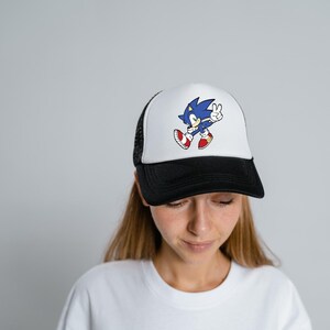 May include: A white and black trucker hat with a blue Sonic the Hedgehog graphic. The graphic shows Sonic running with his arms raised and a peace sign.