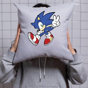 May include: A gray pillow with a blue and red cartoon character of Sonic the Hedgehog running and giving a peace sign. The character is in a white outline.