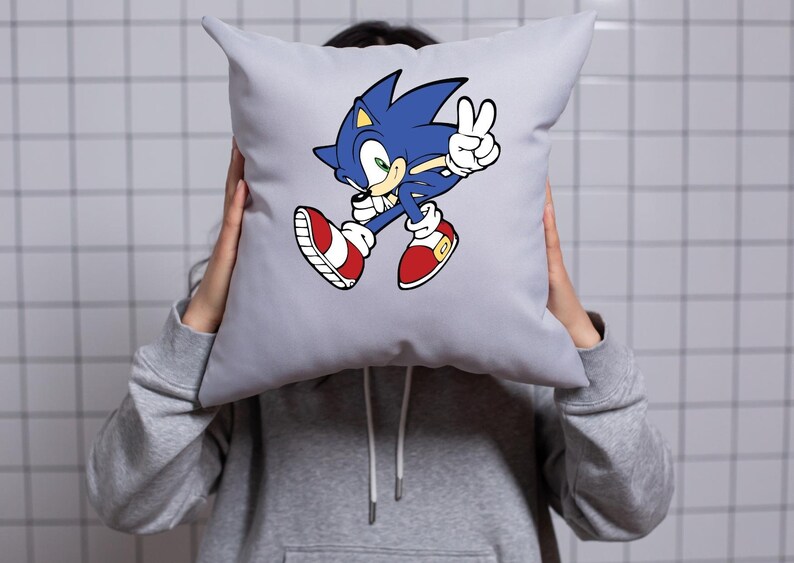 May include: A gray pillow with a blue and red cartoon character of Sonic the Hedgehog running and giving a peace sign. The character is in a white outline.