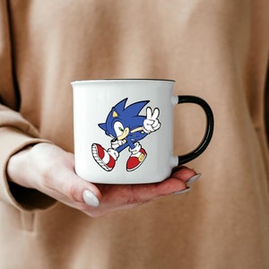 May include: White ceramic mug with a black handle featuring a cartoon image of Sonic the Hedgehog in blue and red running with his hand raised in a peace sign.