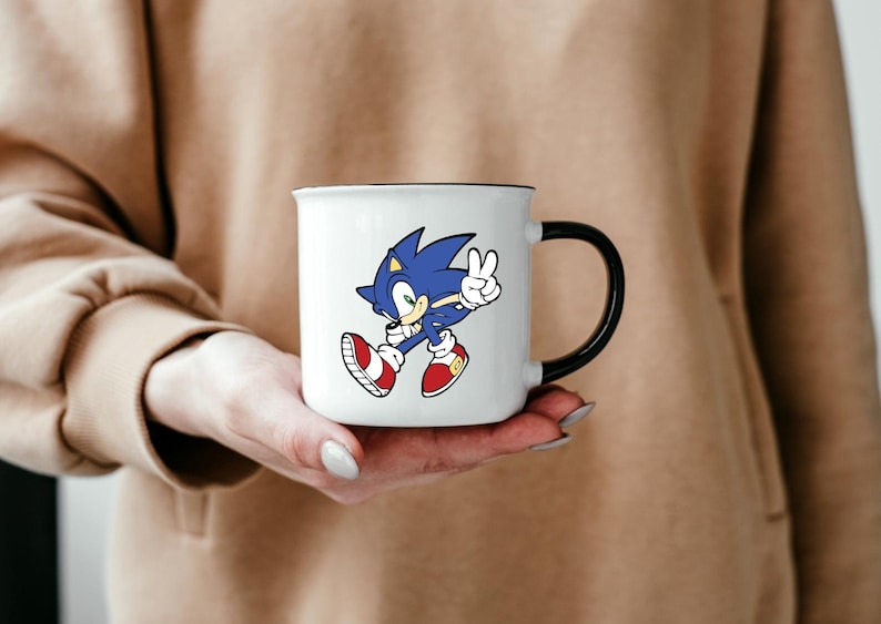 May include: White ceramic mug with a black handle featuring a cartoon image of Sonic the Hedgehog in blue and red running with his hand raised in a peace sign.