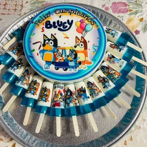 May include: A round cake and several popsicles on a silver platter. The cake features the word "Bluey" and "Feliz Cumpleaños" with cartoon characters. The popsicles have blue and white designs with cartoon characters.