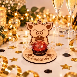 May include: A light-colored wooden stand featuring a laser-cut pig holding a four-leaf clover, with the German text "Viel Gl&uuml;ck" engraved on the circular base. A round red chocolate candy wrapped in clear star-patterned foil sits in the center.