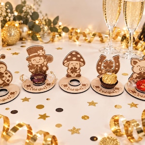 May include: Five light-colored wooden stands featuring laser-cut designs of a ladybug, a person in a top hat, a mushroom, a horseshoe, and a pig. Each stand has a circular cutout for a candy and the German text Viel Gl&uuml;ck engraved on the base.