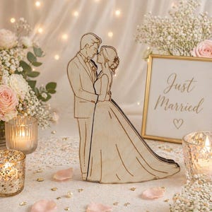 Könnte beinhalten: Ein Kuchenaufsatz aus Holz für eine Hochzeit, der ein sich umarmendes Brautpaar zeigt. Die Braut trägt ein langes Kleid, der Bräutigam einen Anzug. Ein goldgerahmtes Schild mit "Just Married" ist zu sehen. Die Szene umfasst Blumen, Kerzen und Rosenblätter.