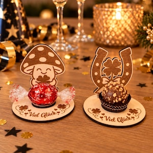 May include: Two circular wooden stands featuring laser-cut designs. One stand displays a smiling mushroom holding a clover, while the other shows a horseshoe with a four-leaf clover. Both bases are engraved with the German text Viel Gl&uuml;ck.
