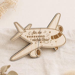 Könnte beinhalten: Ein Holzpuzzle in Form eines Flugzeugs mit dem deutschen Text "für die nächste Reise" (für die nächste Reise). Das Puzzle besteht aus hellem Holz und weist eingravierte Details wie Fenster, Propeller und dekorative Elemente auf. Das Flugzeug ist in zwei Teile geteilt.