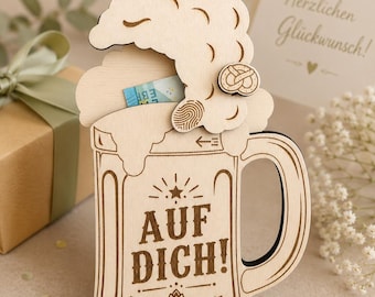 Op de afbeelding: Een lichtgekleurde houten bierpul-uitsparing met een schuimkraag, een klein krakelingdetail en de gegraveerde tekst "AUF DICH!" boven een hopafbeelding. Het ontwerp bevat een gleuf voor het insteken van papiergeld, zichtbaar als een blauwe rand.