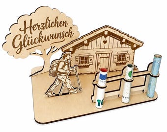 May include: A light-colored wooden display featuring a cutout of a hiker, a cabin, and a tree with the text "Herzlichen Glückwunsch" engraved on it. The base includes a wooden fence with three circular slots holding rolled-up paper currency.