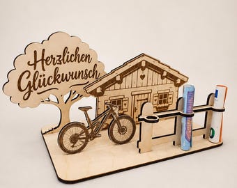 May include: A laser-cut wooden display featuring a tree with the text "Herzlichen Glückwunsch," a small cabin, a mountain bike, and a fence with two cylindrical slots holding rolled-up paper currency on a rectangular wooden base.