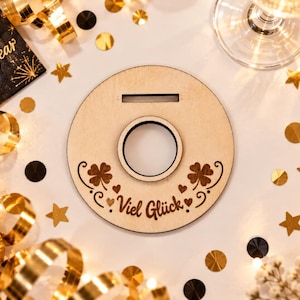 May include: A circular light-colored wooden ornament featuring a central hole, a horizontal slot at the top, and laser-engraved designs of two four-leaf clovers, small hearts, and the German text "Viel Gl&uuml;ck" centered at the bottom of the piece.