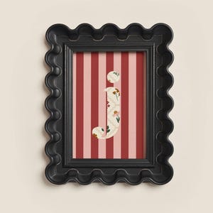 May include: A framed print featuring the lowercase letter "j" with a floral pattern, set against a red and pink striped background. The black frame has a wavy, scalloped edge. The artwork is displayed on a cream-colored wall.