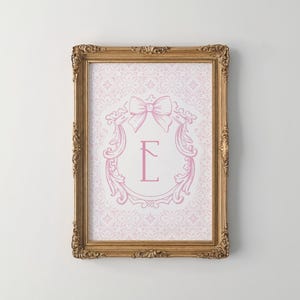Puede incluir: Una impresión enmarcada con una letra monograma rosa "E" dentro de un lazo decorativo y un diseño floral. La impresión está sobre un fondo estampado y enmarcada en un marco dorado ornamentado.