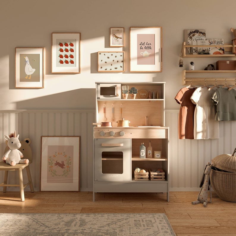 May include: A white play kitchen with a gray oven, a wooden countertop, and shelves. The kitchen has a microwave, a sink, and a stovetop. There are several framed pictures on the wall behind the kitchen, including one that says "Let her be little".