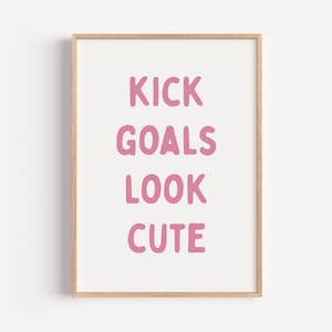 May include: A framed print featuring the phrase "KICK GOALS LOOK CUTE" in pink, handwritten lettering on a white backdrop. The frame is light brown. This print is a motivational piece for a room.