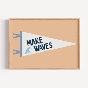 May include: A framed print with a pennant design. The pennant is white with the words "MAKE WAVES" in navy blue and a light blue wave graphic. The background is peach-colored. The frame is made of wood.