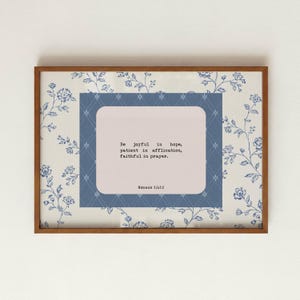 May include: A rectangular wall art print in a wooden frame featuring a cream background with a blue floral pattern. A central blue rectangle with a white diamond grid contains a light pink box with the text: Be joyful in hope, patient in affliction, faithful in prayer. Romans 12:12.