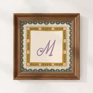 May include: A square wooden frame containing a print with a central lavender letter M on a cream background. The border features a mustard yellow square with green leafy vines and small pink flowers, surrounded by a dark teal scalloped edge.