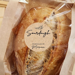Könnte beinhalten: Ein Sauerteigbrot in einer transparenten Plastiktüte, in einer braunen Papiertüte. Die Tüte hat einen runden Aufkleber mit der Aufschrift "Sourdough Homemade by Marian". Das Brot liegt auf einem Holzschneidebrett.