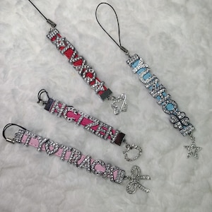 May include: Four phone charms with different band names. The charms are made of silver metal with rhinestones and have a black string attached. The charms spell out the names "NCTZEN", "ITZY", "TWICE", and "YUNHO".