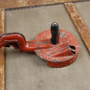 May include: A red metal tool with a black handle and a clamp. The tool has a round base with a hole in the center. It is used for making holes in metal.