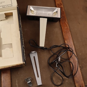 May include: A black and silver light with a white stand and a black cord. The light is in a box with a white interior and the words "Made in Denmark" printed on the box.