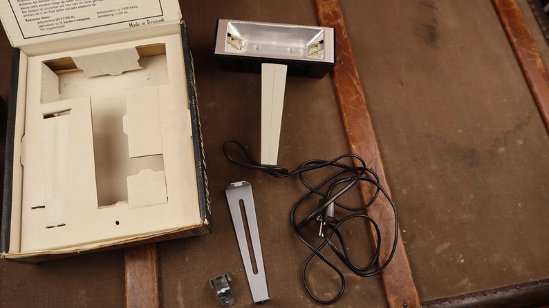 May include: A black and silver light with a white stand and a black cord. The light is in a box with a white interior and the words "Made in Denmark" printed on the box.