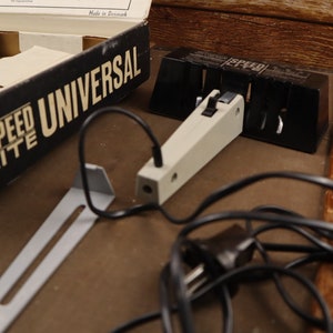 May include: A black and white box with the text "Speed Lite Universal" on it. The box is open and there is a white and black flash unit with a black cord attached to it. There is also a metal bracket and a black plug.