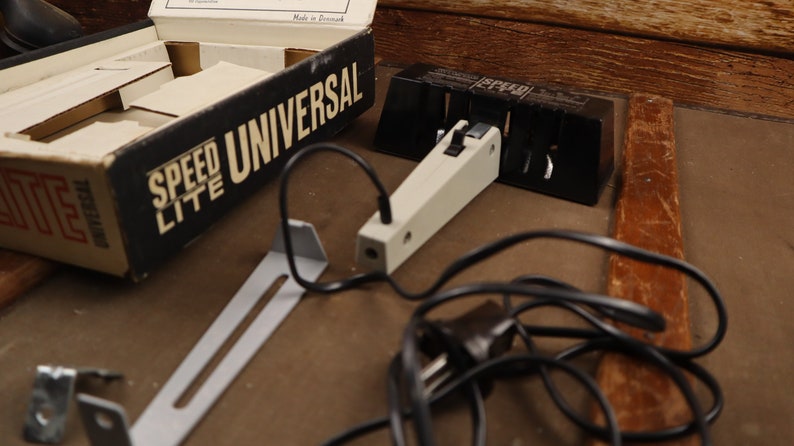 May include: A black and white box with the text "Speed Lite Universal" on it. The box is open and there is a white and black flash unit with a black cord attached to it. There is also a metal bracket and a black plug.