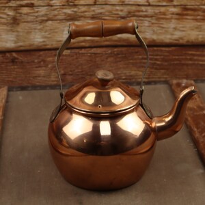 May include: A copper tea kettle with a wooden handle and spout. The kettle has a round body and a lid with a small wooden knob. The handle is made of wood and metal. The kettle is likely vintage or antique.