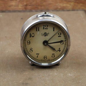 May include: A silver alarm clock with a white face and black hands. The clock is set to 3:45. The clock is on a brown surface.