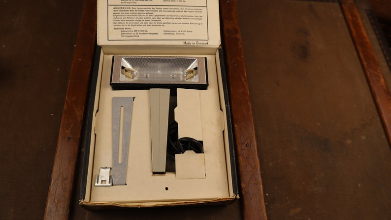 May include: A white box with a black lid containing a silver and black light fixture with a white cord. The box is labelled "Made in Denmark".
