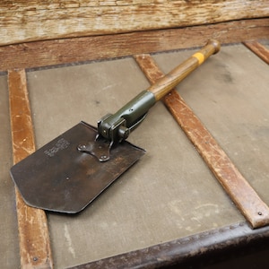 Könnte beinhalten: Eine klappbare Schaufel mit einem Holzstiel und einer Metallklinge. Die Schaufel liegt auf einer Holzoberfläche.