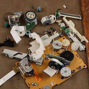 Può includere: Primo piano di un lettore CD o DVD smontato. I pezzi sono sparsi su una superficie marrone, tra cui una scheda del circuito, ingranaggi, motori e una lente laser.
