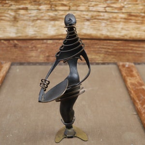 Puede incluir: Una escultura de metal negro de una persona sosteniendo un objeto redondo. La escultura está hecha de metal retorcido y tiene una base dorada.