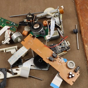 Op de afbeelding: Een verzameling gedemonteerde elektronische componenten, waaronder printplaten, tandwielen, motoren en draden, verspreid over een bruin oppervlak.