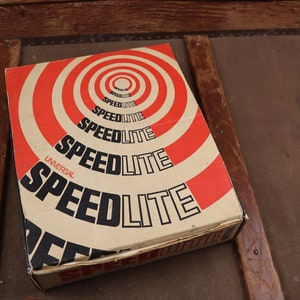 May include: A vintage cardboard box with a red and white target design. The box is labelled "Universal Speedlite" in black lettering.
