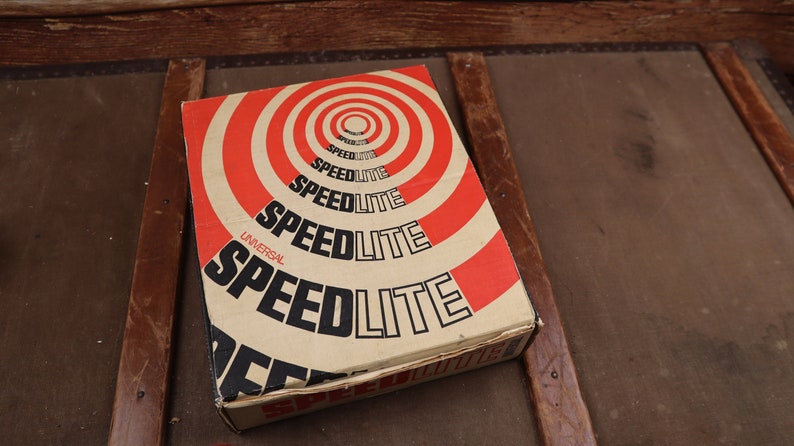May include: A vintage cardboard box with a red and white target design. The box is labelled "Universal Speedlite" in black lettering.