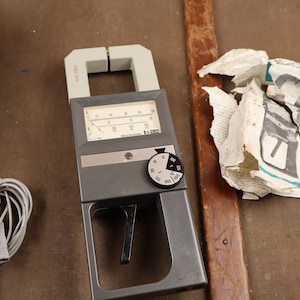 Puede incluir: Un medidor de pinza gris con un dial y una perilla negra. El medidor tiene una cara blanca con marcas negras. El medidor de pinza está sobre una superficie marrón con una caja de cuero negra y un cable gris enrollado.