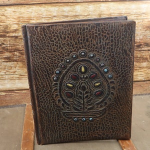 Puede incluir: Un diario encuadernado en cuero marrón con un diseño decorativo en relieve que representa un árbol con piedras preciosas de colores. El diario tiene una superficie texturizada y un aspecto desgastado.