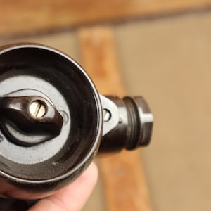 Op de afbeelding: Een zwarte bakelieten lichtschakelaar met een ronde knop en een zilveren schroef. De schakelaar is gemarkeerd met "OM 25".